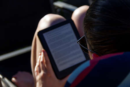 Brecht, Belgium - May 8 2018: A close up portrait of a person reading an e-book on an e-ink e-reader on a sunny day outside in the garden. An easy way to travel with a pocket sized library.のeditorial素材