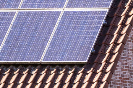 A close up portrait of solar panels lying on an orange tile roof. The photovoltaic cells of the...
