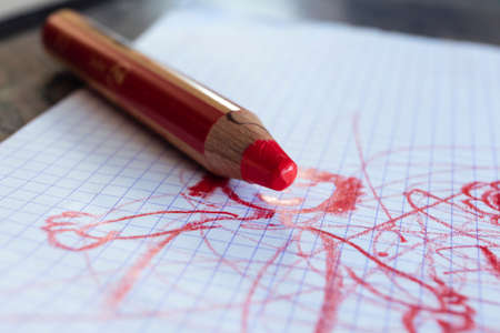 A portrait of a large thick red coloring pencil for small children on a white checkered sheet or page with a cute toddler drawing or doodle on it.の写真素材