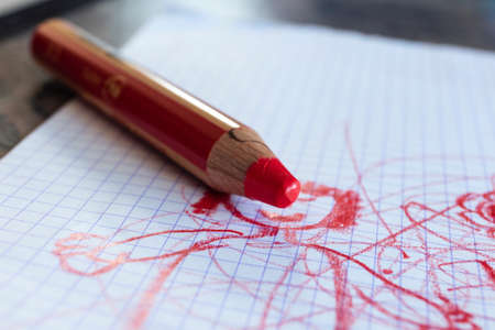 A close up portrait of a large thick red coloring pencil for small children on a white checkered sheet or page with a cute toddler drawing or doodle on it.