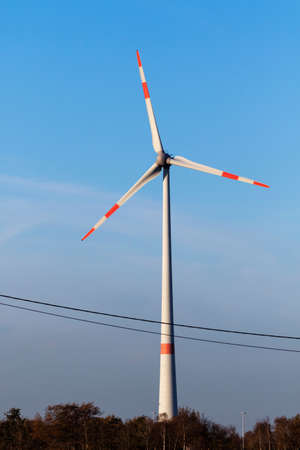 A portrait of a big windmill standing tall above the tree line. The turbine generates clean...