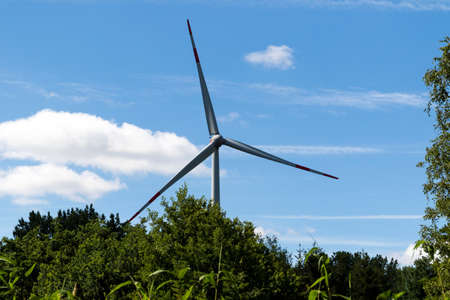 Horizontal portrait of a windmill standing high above the trees in a blue sky, generating sustainable green ecologic electric energy by catching wind on its wings and turning around using a generator.の写真素材
