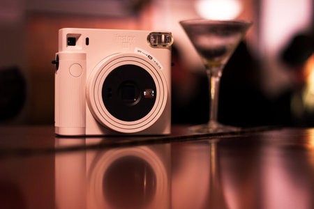 Mechelen, Belgium - December 10, 2022: A portrait of a polaroid instax photo camera standing on a wooden surface with a cocktail glass next to it and their mirrored reflection at a social party.のeditorial素材