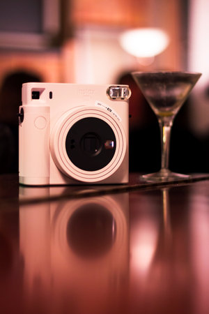Mechelen, Belgium - December 10, 2022: An instax polaroid photo camera standing on a wooden surface with a cocktail glass next to it and their mirrored reflection at a social party.のeditorial素材