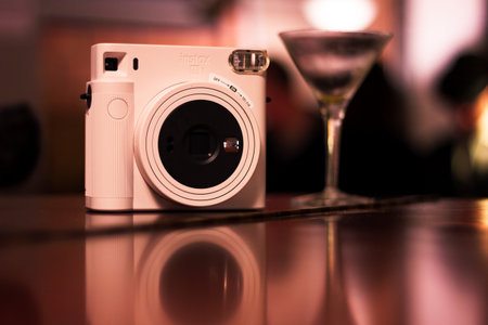 Mechelen, Belgium - December 10, 2022: A close up of a polaroid instax photo camera standing on a wooden surface with a cocktail glass next to it and their mirrored reflection at a social party.のeditorial素材