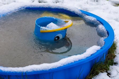 A portrait of a blue bucket with a face painted on it and a yellow handle for a child to play with lying in a blue play shell pool which is frozen and surrounded by snow in the winter.の写真素材