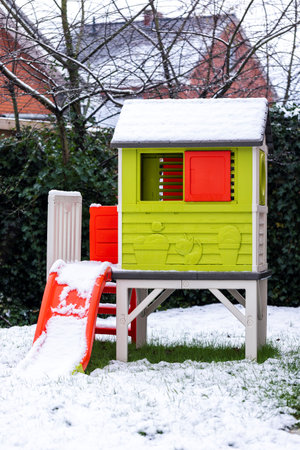 A vertical portrait of a tiny green play house with a red slide standing in a garden covered in snow during winter. The house has tiny red windows and a door and is ready to be played with.の写真素材