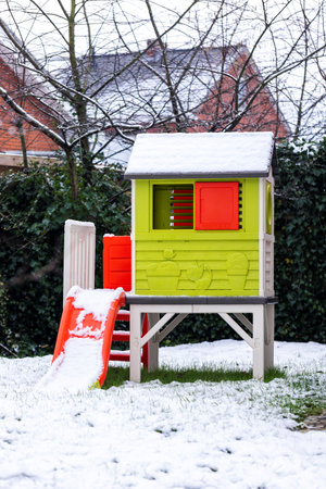 A portrait of a green small play house with a red slide standing in a garden covered in snow during winter. The house has tiny red windows and a door and is ready to be played with.の写真素材