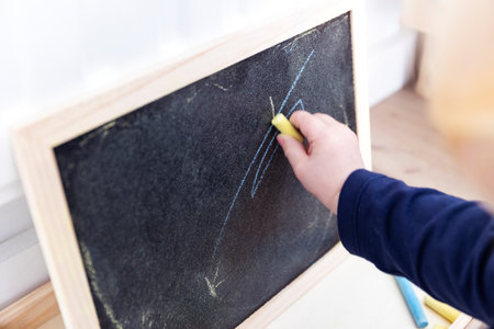 A close up portrait of a toddler drawing, doodling or scribbling a bit on a chalkboard. The child is writing on the blackboard and has a yellow piece of chalk in its hand.の写真素材