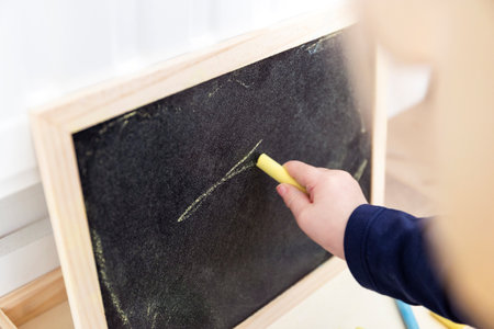 A close up portrait of a toddler scribbling, doodling or drawing a bit on a chalkboard. The child is writing on the blackboard and has a yellow piece of chalk in its hand.の写真素材