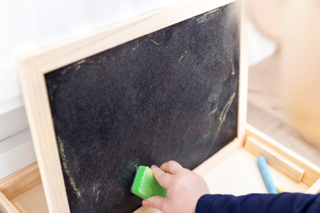 A close up portrait of a toddler drawing, scribbling or doodling a bit on a chalkboard. The child is wiping the blackboard and has a green sweeper in its hand.の写真素材