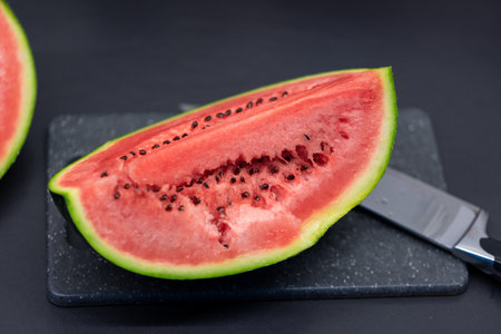 A portrait of a quarter cut of a green watermelon lying on a plastic cutting board next to a knife. The piece or slice of the fruit is ready to be eaten and you can see black seeds in the red pulp.の写真素材