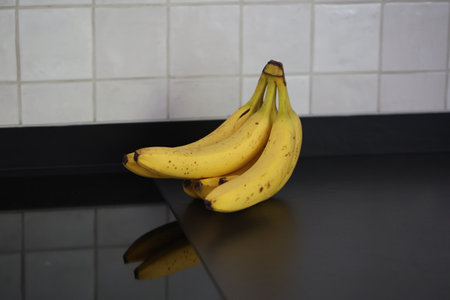 A portrait of a bunch of yellow bananas with som brown spots lying on a black kitchen countertop. The delicious energizing food is ready to eat and ideal before a sport activity.の写真素材