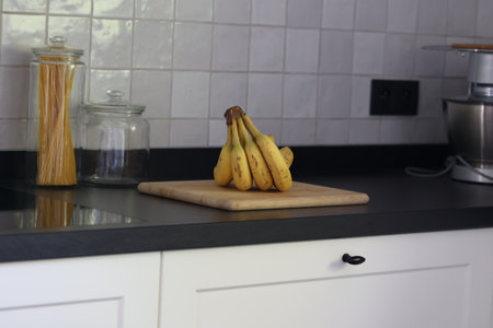 A portrait of a bunch of yellow bananas lying on a wooden cutting board on a black kitchen countertop. The delicious energizing food is ready to eat and ideal before sport.の写真素材