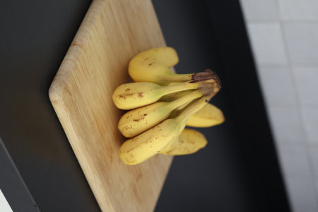 A portrait of a bunch of yellow bananas lying on a wooden cutting board on a black kitchen countertop. The delicious energizing food is ready to eat and ideal before sport.の写真素材