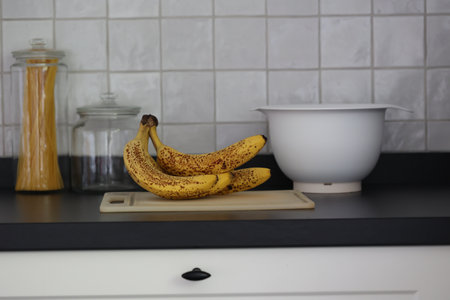 A portrait of a bunch of yellow bananas lying on a white plastic cutting board on a black kitchen countertop. The delicious energizing food is ready to eat and ideal before sport.の写真素材