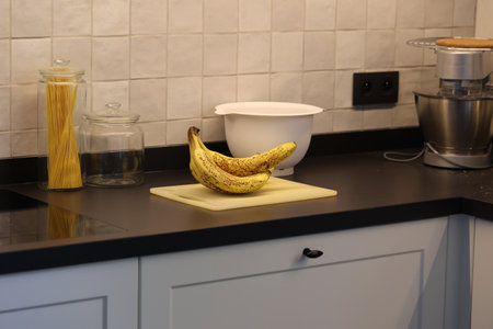 A portrait of a bunch of yellow bananas lying on a white plastic cutting board on a black kitchen countertop. The delicious energizing food is ready to eat and ideal before sport.の写真素材