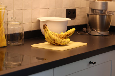 A portrait of a bunch of yellow bananas with brown spots lying on a white plastic cutting board on a black kitchen countertop. The delicious energizing food is ready to eat and ideal before sport.の写真素材