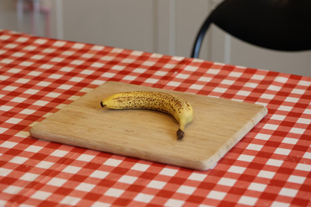 A portrait of a single yellow banana with some brown spots lying on a wooden cutting board on a red and white table cloth on a kitchen table. The delicious energizing food is ready to be eaten.の写真素材