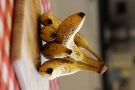 A vertical of yellow bananas with some brown spots lying on a wooden cutting board on a red and white table cloth on a kitchen table. The delicious energizing food is ready to be eaten.の写真素材