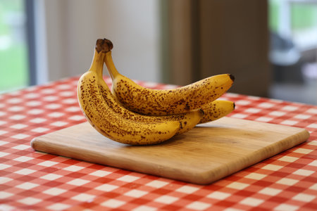 A portrait of yellow bananas with some brown spots lying on a wooden cutting board on a red and white table cloth on a kitchen table. The delicious energizing food is ready to be eaten.の写真素材