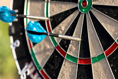A closeup of a part of a dartboard game standing outdoors on a sunny day, with two darts sticking in...