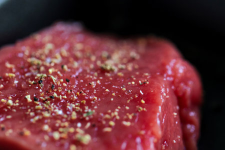 A close up portrait of the seasoning on a red raw steak being prepared or cooked in a frying pan. The delicious meat has salt and pepper on it for a perfect dinner or meal.の写真素材