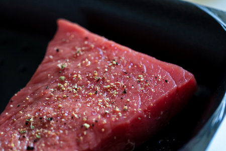 A close up portrait of the seasoning on a red raw steak being prepared or cooked in a frying pan. The delicious meat has pepper and salt on it for a perfect dinner or meal.の写真素材