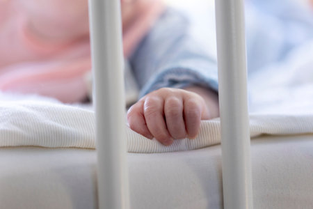 A close up portrait of only the small hand of a cute baby lying on a cushion between the bars of a baby box. The little fingers are grasping a sheet.の写真素材
