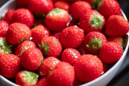 A close-up portrait of fresh, ripe strawberries with vivid red color and green leaves in a white bowl, showcasing their juicy texture and natural beauty. The delicious fruit is ready to eat.の写真素材