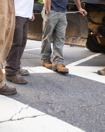 Four men, cropped from the waist down, wearing workmen's pants and boots stand around a piece of construction equipment on a road.の写真素材