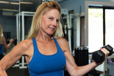 Female middle age fitness model shows muscular and lean arm strength while performing a dumbbell curl in gym.のeditorial素材