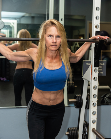 Serious female weight lifted showing strong abdominal muscles while posed in gym squat rack.のeditorial素材