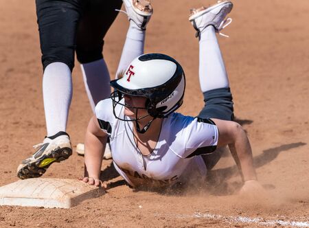 Softball player in white uniform diving back to first base in cloud of dust during game between...