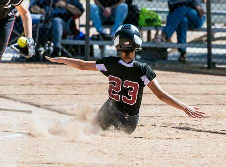 Female teenage softball player in black uniform sliding into home plate before the catcher can make the tag.の写真素材