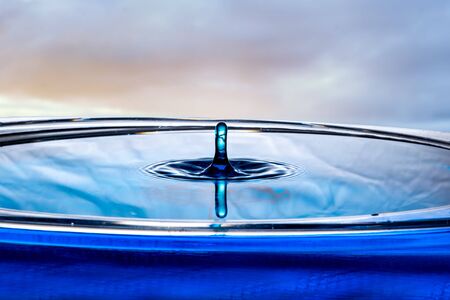 Single drop of water rises out of a calm pool surface into a rounded top column.