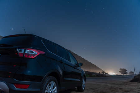 Black compact car parked along highway as constellation Orion is overhead in the star lit night sky.の写真素材