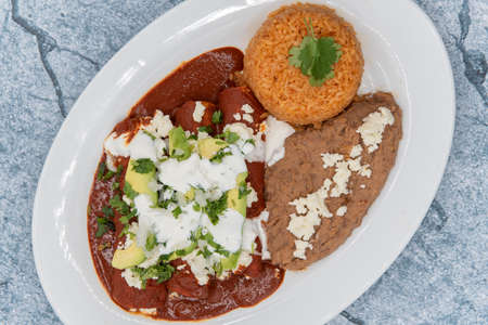 Overhead view of chicken enchiladas smothered in red sauce served with refried beans and rice for a...