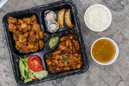 Overhead view of bento box packed to go loaded with sesame chicken, spicy chicken, salad, gyoza dumplings, rice, and miso soup for a complete take out meal.の写真素材