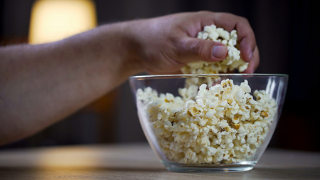 Closeup of male hand taking popcorn from bowl on table, unhealthy food addictの写真素材
