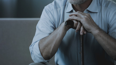 Close-up of adult male hands holding walking stick, old man sitting on sofaの写真素材