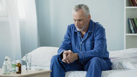 Upset ill senior male sitting on bed, taking medication to recover from illnessの写真素材