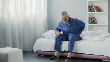 Retired man sitting in bed and feeling terrible pain in back, health and illnessの写真素材