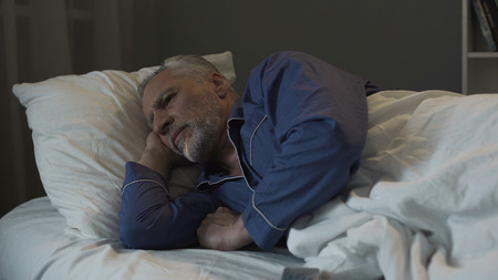 Gray haired man lying in bed and suffering from insomnia, depression and healthの写真素材