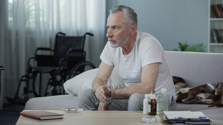 Old handicapped male sitting on couch and taking pills, loneliness and sadnessの写真素材
