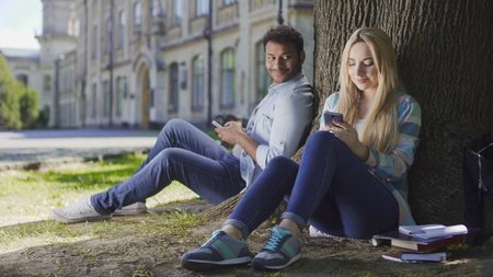 Man with cellphone sitting under tree and looking at girl using phone, affectionの写真素材