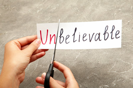 Scissors cutting white paper with the text unbelivable, change word to belivable. Hand holding card with the text unbelievable. Top viewの写真素材