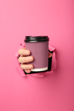 Female hand with cup of coffee punching through the pink paper. top viewの写真素材