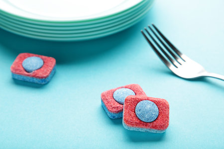 Dishwasher detergent tablet with fork and dishes on blue background. top view