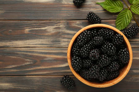Ripe blackberries with leaves in a wooden bowl on brown wooden background. top view.